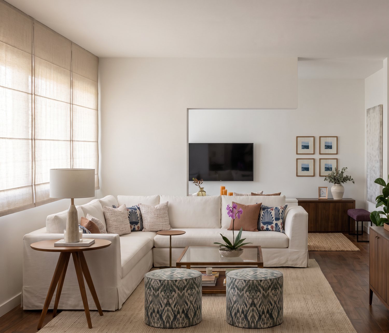 Sala com sofá de canto claro, pufes estampados e mesa de centro de vidro